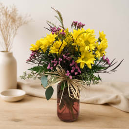 Yellow daisies and pink filler flowers in a glass vase with a raffia bow