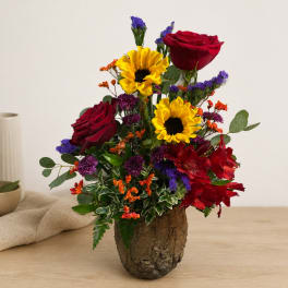 Mixed bouquet with red roses, yellow sunflowers, and purple accents in a rustic vase
