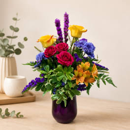 Colorful bouquet of roses, purple spikes, and blue flowers in a purple vase