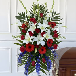 Tall funeral spray with white lilies, red gerberas, and blue flowers beside a casket
