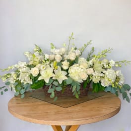 Low rectangular floral arrangement with white lilies, roses, and orchids in a woven basket.