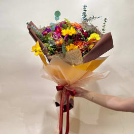 Handheld bouquet of mixed bright flowers wrapped in brown and peach paper