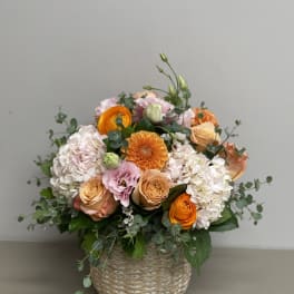Round arrangement of peach and orange roses, hydrangeas, and pink blooms in a woven basket