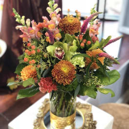 Tall mixed arrangement with orange mums, snapdragons, and green orchid in a glass vase with gold band