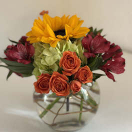 Mixed bouquet with orange roses, yellow sunflower, green hydrangea, and burgundy flowers in a glass vase