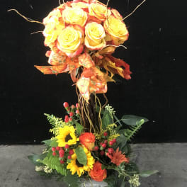 Tall floral arrangement with yellow and orange roses, sunflowers, and mixed greenery in a vase.