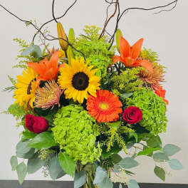 Bright mixed bouquet in a clear glass vase with sunflowers, lilies, and orange blooms