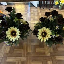 Two floral arrangements with yellow sunflowers and dark roses in glass vases