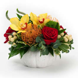 Low arrangement of yellow orchids, orange protea, and red roses in a white pumpkin-shaped bowl