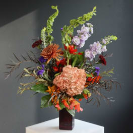 Tall mixed flower arrangement in a dark vase with peach, purple, and orange blooms