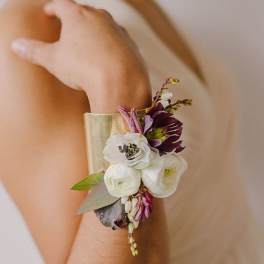 Gold cuff bracelet corsage with white and plum flowers worn on a person's forearm