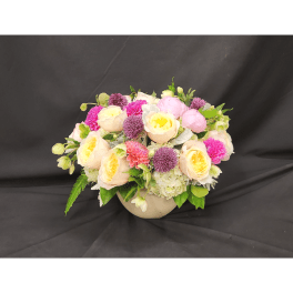 Round bouquet of pale roses, pink blooms, and purple flowers in a beige vase