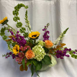 Mixed bouquet with sunflowers, peach roses, and purple blooms in a pale green pumpkin vase