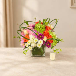 Colorful mixed flower arrangement in a square glass vase with a candle beside it