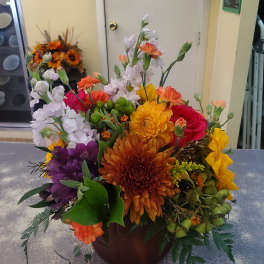 Mixed bouquet in a brown container with orange, yellow, purple, pink, and white blooms.