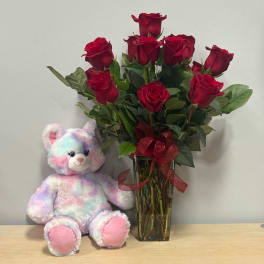 Red roses in a glass vase beside a pastel teddy bear