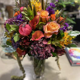Tall mixed arrangement of bright roses, calla lilies, hydrangeas, and accent blooms in a clear glass vase