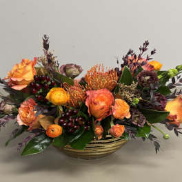 Low centerpiece of orange roses, pincushion protea, and dark berries in a striped gold bowl