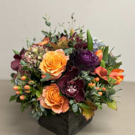 Low arrangement of orange roses, dark burgundy orchids, and mixed blooms in a square dark wooden vase