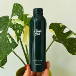 Hand holding a dark green plant fertilizer bottle in front of variegated monstera leaves