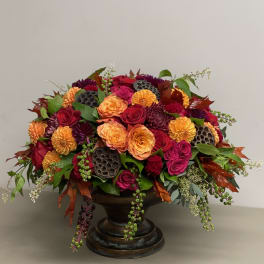 Low arrangement of orange and red roses with dark accent flowers in a bronze pedestal bowl