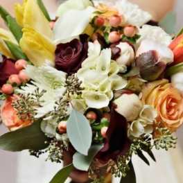 Hand-tied bouquet of ivory, peach, yellow, and burgundy flowers with berries, held by a person in white.