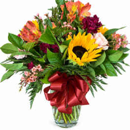 Mixed bouquet with sunflowers, alstroemeria, carnations, and a red ribbon in a glass vase