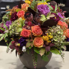 Lush autumn-toned arrangement with orange roses, protea, orchids and mixed blooms in a round ceramic vase