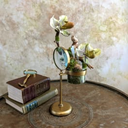 Small floral arrangement with striped blooms in a glass container beside a magnifying glass and books