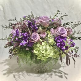 Low pedestal arrangement of lavender roses, purple blooms, and green hydrangeas in a glass compote vase
