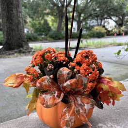 Orange flowers arranged in a pumpkin-shaped container with a fall ribbon