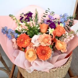Mixed bouquet of orange, purple, and blue flowers wrapped in pink paper