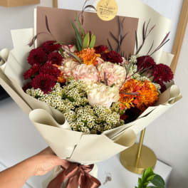Handheld bouquet of burgundy, cream, and orange flowers wrapped in beige paper