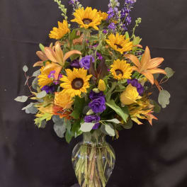 Mixed bouquet of sunflowers, lilies, and purple blooms in a clear glass vase