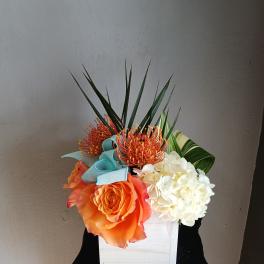 Orange rose and white hydrangea arrangement in a white box with blue ribbon