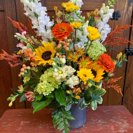 Mixed bouquet with sunflowers, orange zinnias, white snapdragons, and green hydrangeas in a metal vase