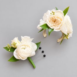 Two cream floral boutonnieres with green leaves on a gray background
