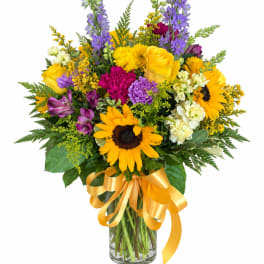 Mixed bouquet of sunflowers, roses, and purple flowers in a glass vase with a yellow ribbon