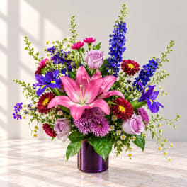 Colorful bouquet with pink lilies, lavender roses, and blue flowers in a purple vase