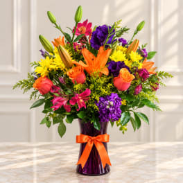 Colorful bouquet of lilies, roses, and daisies in a purple vase with an orange ribbon