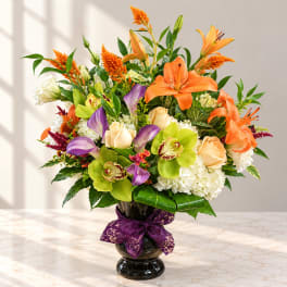 Orange lilies and purple orchids in a dark glass vase with a purple ribbon