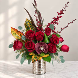 Red roses arranged in a silver vase with burgundy orchids and patterned leaves