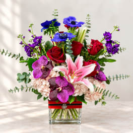 Mixed bouquet of red roses, pink lilies, purple orchids, and blue anemones in a glass vase