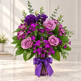 Purple roses and mixed blooms in a glass vase with a purple ribbon