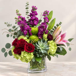 Mixed bouquet of red roses, purple blooms, lilies, and green hydrangeas in a glass vase
