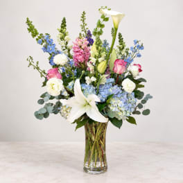 Tall mixed bouquet in a clear glass vase with lilies, roses, hydrangeas, and blue flowers