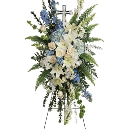 Standing floral spray with white lilies, cream roses, and blue flowers around a cross