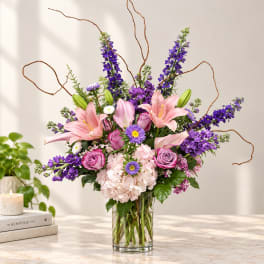 Pink lilies and purple flowers arranged in a clear glass vase