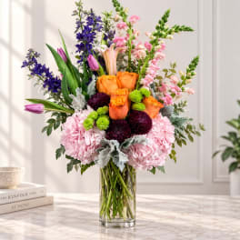 Mixed bouquet of orange roses, pink hydrangeas, and purple flowers in a clear glass vase