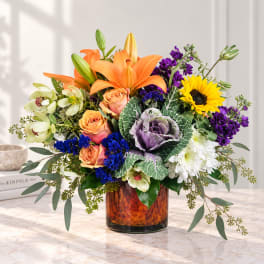 Mixed bouquet with orange lilies, roses, sunflowers, and purple flowers in a glass vase